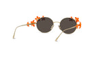 A pair of round sunglasses with a gold frame and dark lenses, adorned with orange flower-shaped embellishments on the sides.